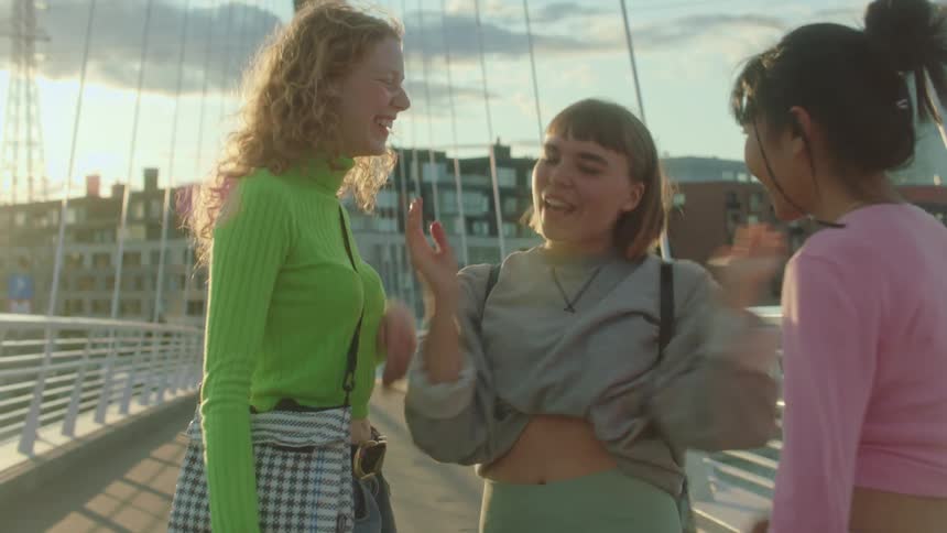 Three woman on a bridge hugging with buildings the sky and clouds in the background