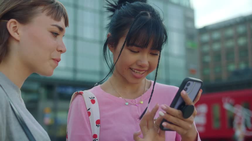 Two young women laugh as they look at a smartphone while standing outside downtown