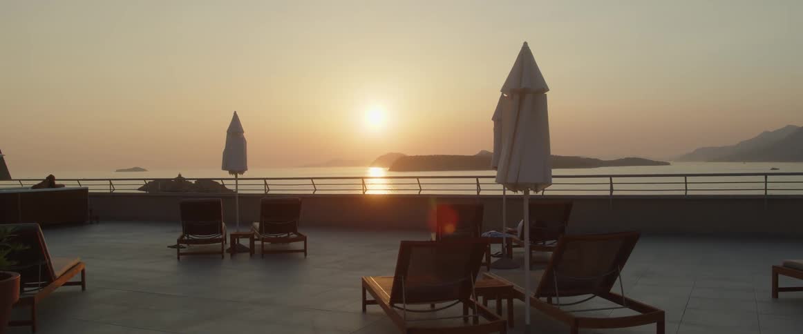Person sits in wooden lounge chair in outdoor seating area near rail overlooking ocean reflecting sun sitting in red sky above distant opposing shore on a misty day