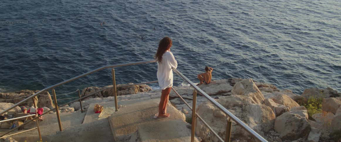 Woman stands near turn of staircase leading to beach with man sitting barechested on shore and both look over the ocean