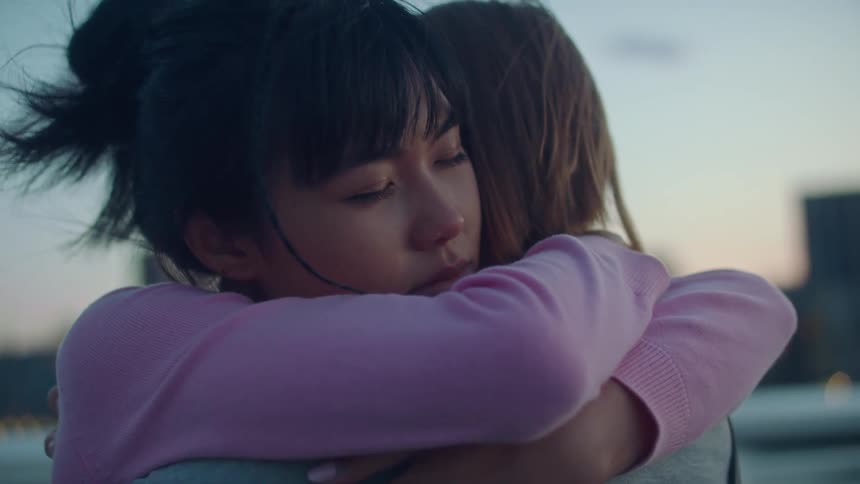 Woman looks into another womans downcast expression and hugs them with a slight smile as red tinted sky and buildings sit in background