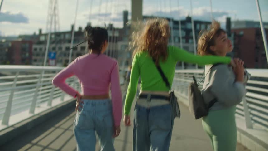 Three girls are walking across a bridge with their arms around each other and holding hands