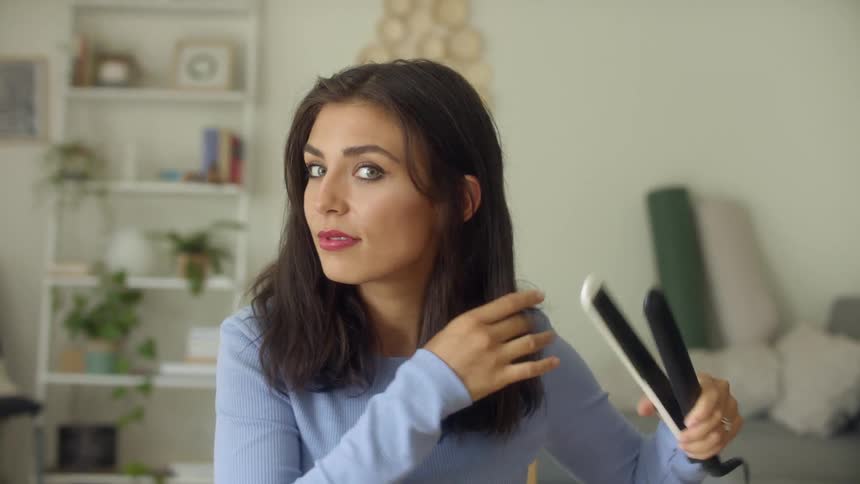 A woman with a flat iron straightening her hair in a room with a wall and a shelf in the background