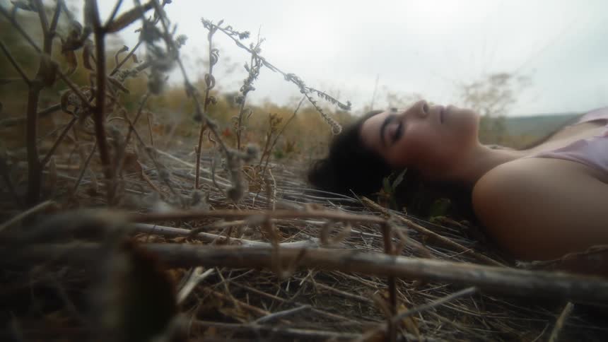 A woman is lying on the ground among plants and sticks and she turns her head to look at the camera