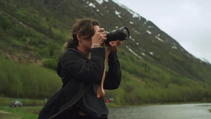 Female photographer stands on shore focusing camera as car drives along road toward mountain behind her