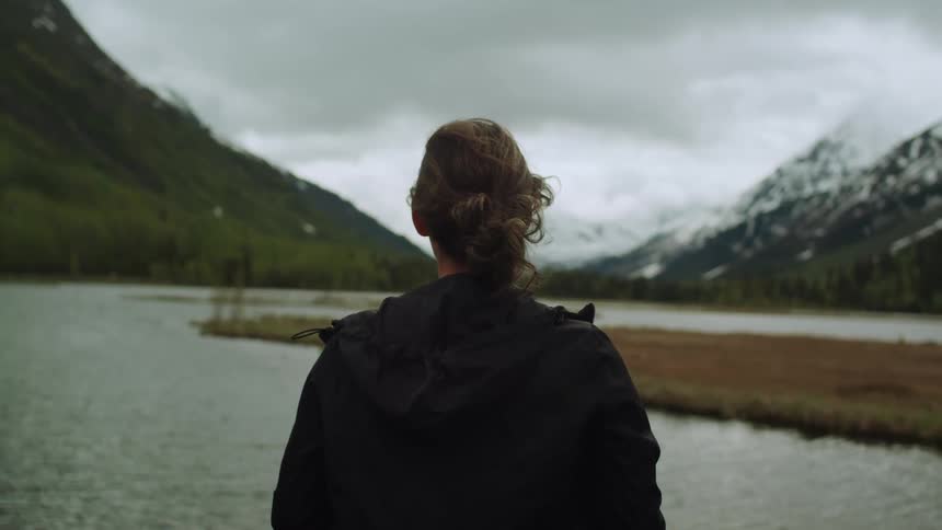 A woman standing on the shore looks out over a river winding through a mountain valley on a cloudy day