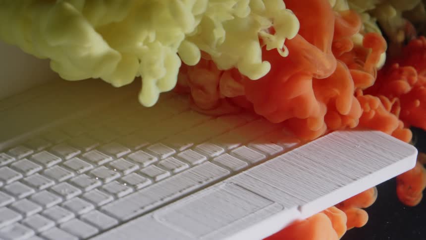 A 3d printed laptop computer has a cloud of orange and yellow smoke blown at it and billow around it