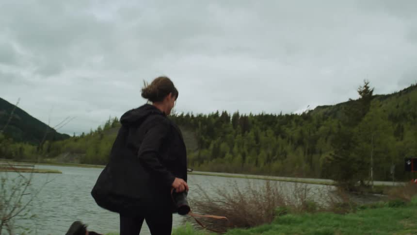 A woman carrying a camera runs on a dirt road beside a lake with hills and trees