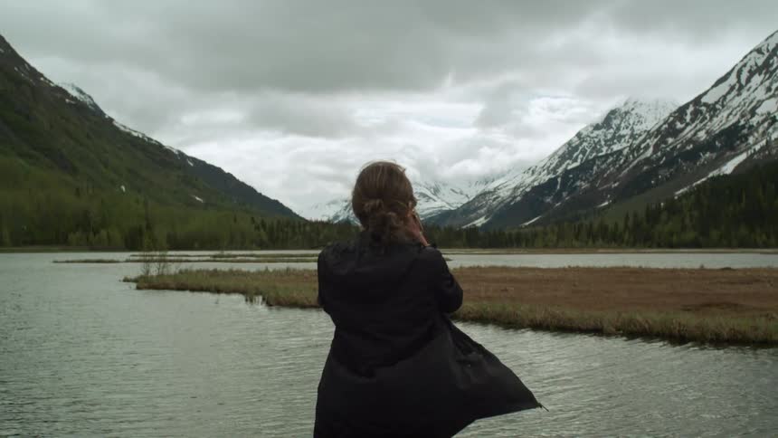 A woman standing with a lake a mountain range the sky and clouds in the background