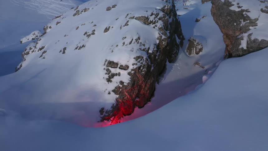 Red glow colors nearby snow covered rock outcropping as smoke flows across mountain slope
