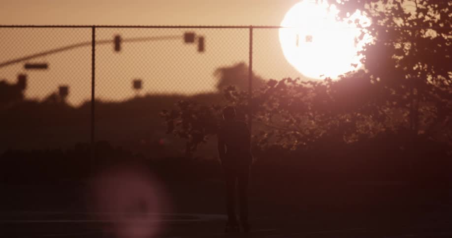 The sun is setting behind a chain link fence in an orange sky as someone rides a skateboard