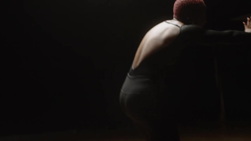 A woman in a black leotard is performing modern dance in a shadowy dance studio