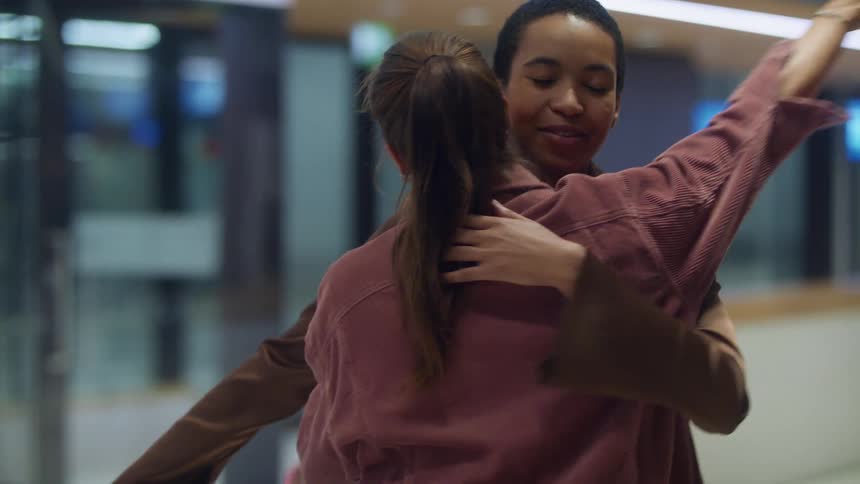 Woman greets arriving friend at terminal as transit passes by outside glass doors
