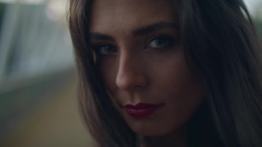 A young woman standing outdoors near a bridge is staring into the camera at dusk