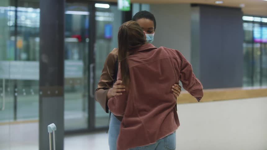 Woman with black mask and ponytail and woman with backpack brown shirt and jeans cuddle each other at the airport