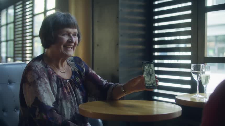 Elderly woman sitting at the table on a restaurant shows vintage pictures on a smartphone