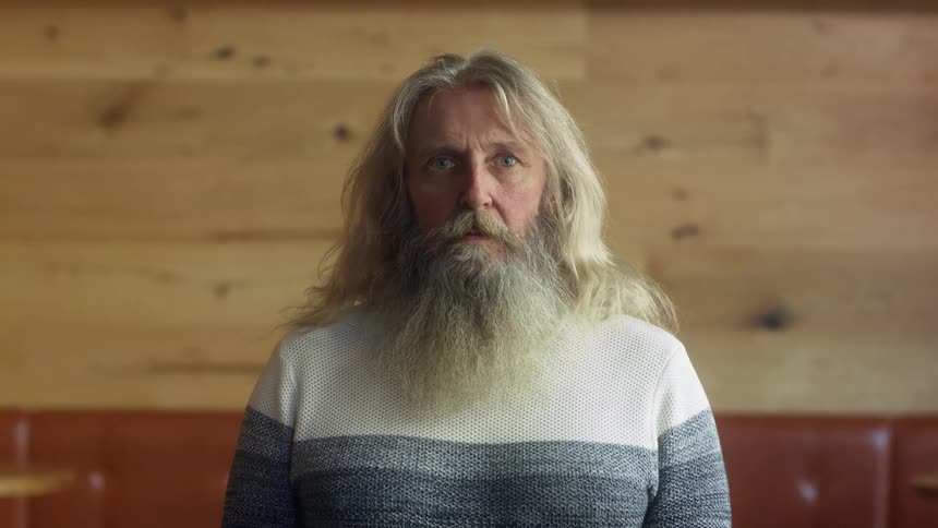 A bearded middle aged man stares into the camera while standing indoors