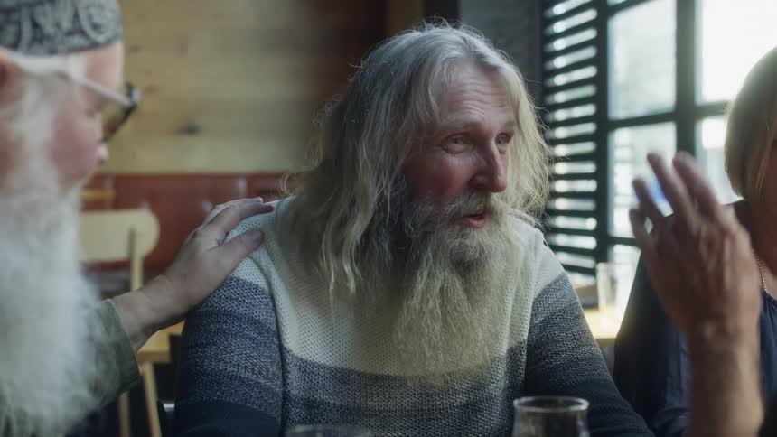 A group of elderly men with long hair and grey beards sits at table having a conversation in pub