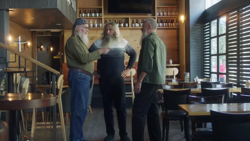 A group of graying men talk while standing together in a restaurant during the day