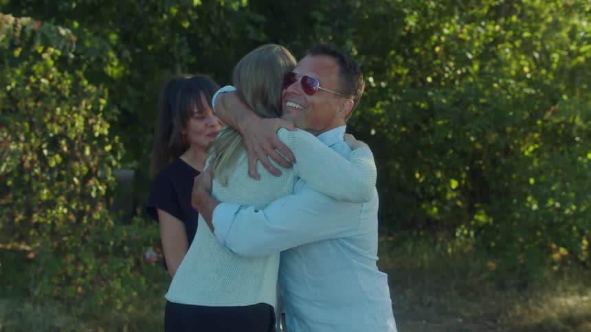 A couple greets a woman with a hug before beginning to chat while standing together outdoors