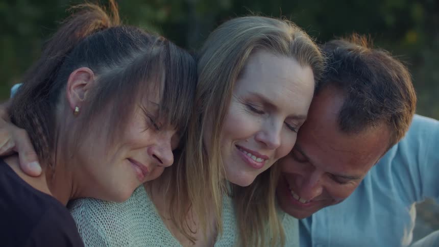 Three people hug each other while sitting outside in the grass at dusk