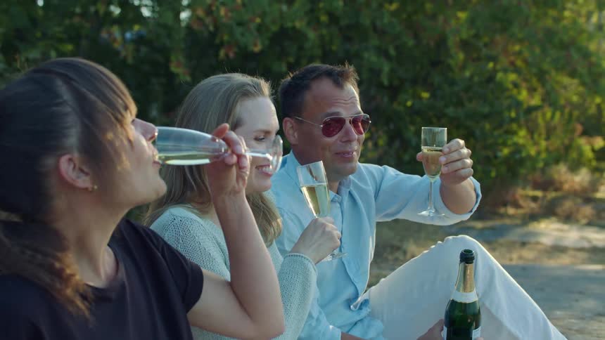 Friends sit on ground near trees drinking wine