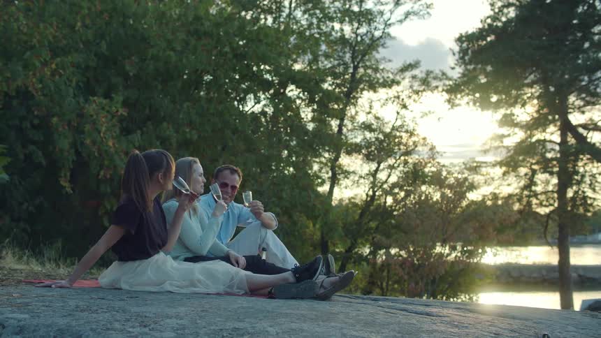 Two women and a man drink wine together while sitting on the stone shore of a river during the day