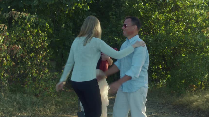 Man with glasses and blue shirt and women wearing skirt black shirt blouse and pants cuddle each other on the dirt road in front of trees