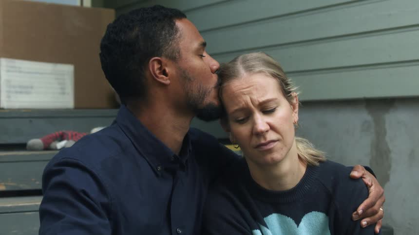 Man consoling a sad woman with a hug and kiss on the forehead while sitting on the stairs of the porch