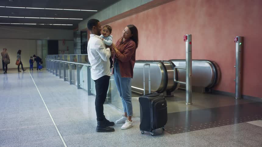 A man holding an infant happily embraces a woman who just stepped off the nearby escalator