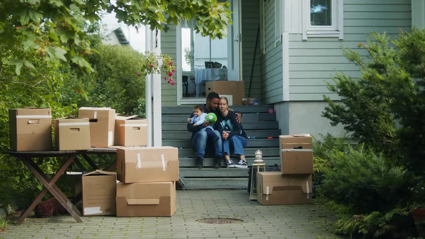 Man carries baby out of house and down steps to sit with wife looking sad in front of several packed boxes