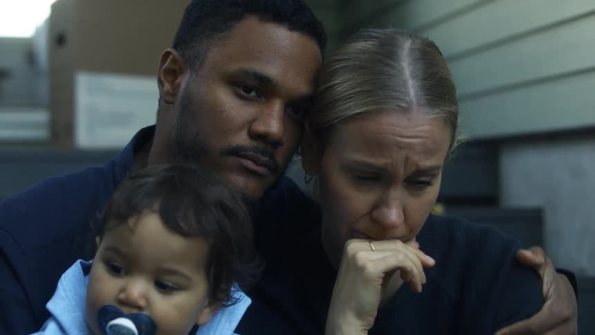 A man embraces and kisses a crying woman as they sit together outdoors while holding a child