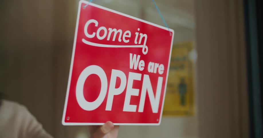 Female staff flips closed sign to show open with signage discussing social distancing in the background
