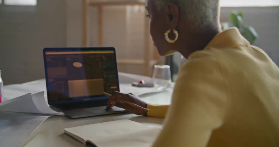 A woman in a yellow shirt talking while working on a laptop in an office setting