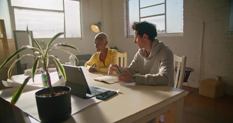 Two coworkers sitting at a desk in an office talking in front of laptops