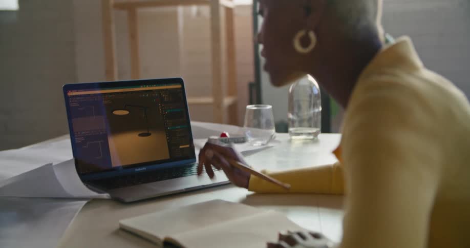 Woman with short grey hair earrings and yellow sweatshirt uses laptop and talks to person in office