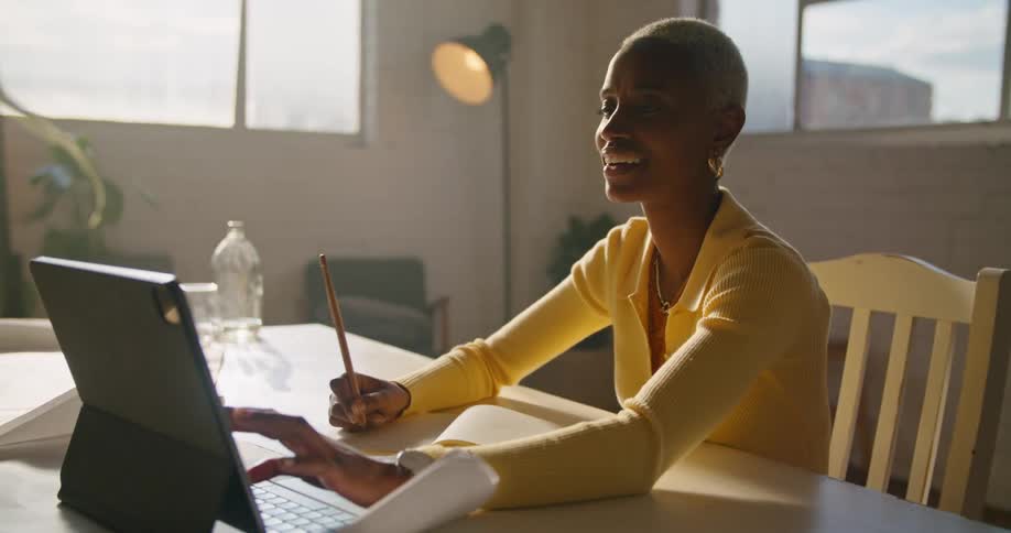 Woman with short hair hoop earrings and yellow sweater sits at table and uses tablet making video call