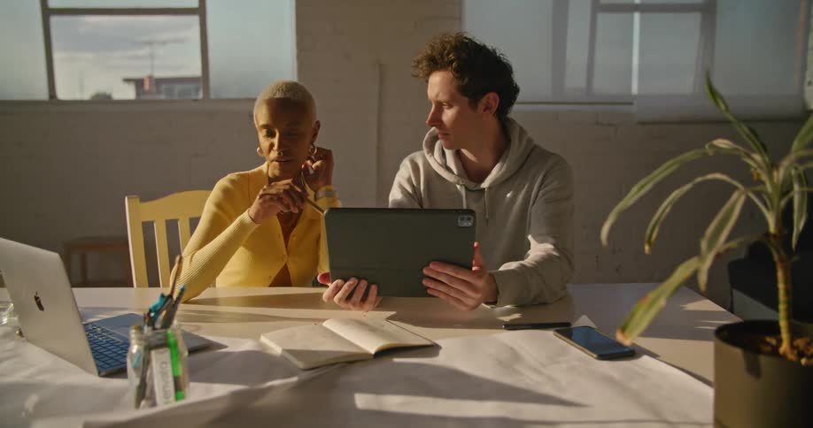 Woman with short hair hoop earrings and yellow sweater sits at table with man wearing grey hoodie using tablet and talking with each other in the office