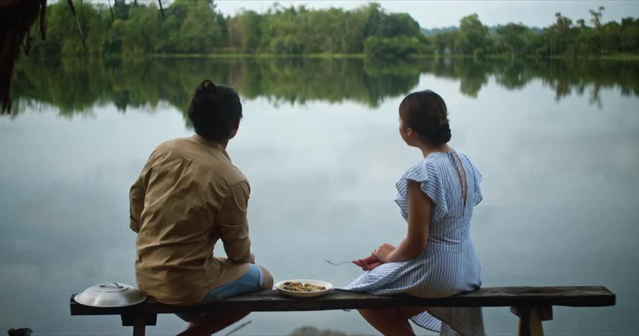 A man and a woman sitting by a lake with trees the sky and clouds in the background