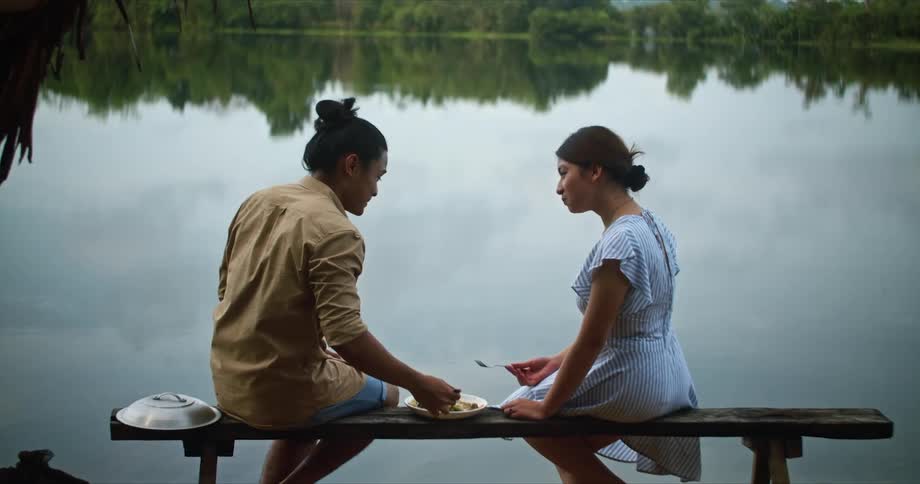 A man and woman are eating food while sitting on abench on the edge of a lake