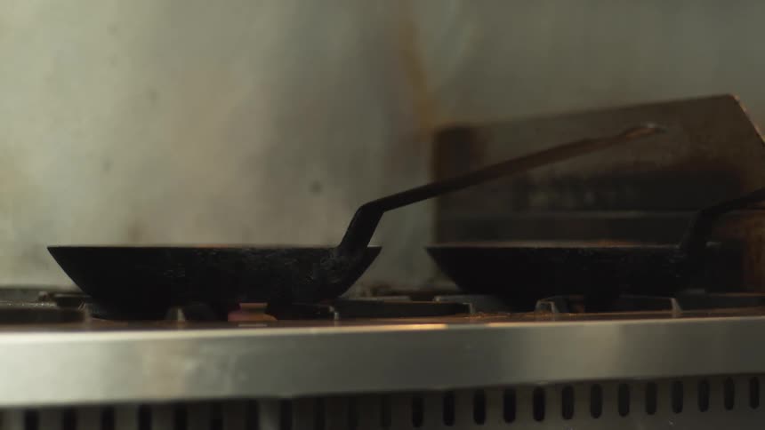 A person adding a liquid to a frying pan on a stove with a flame with a wall in the background