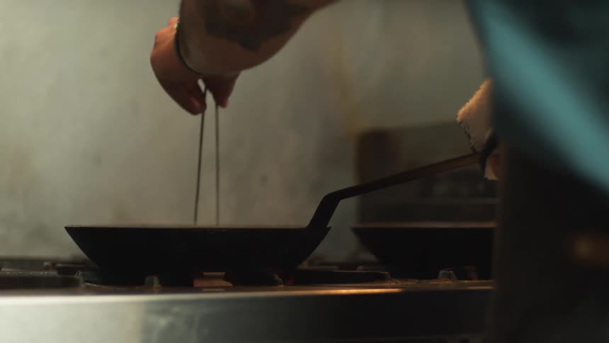 A man is cooking on a gas stove top using a skillet and chopsticks