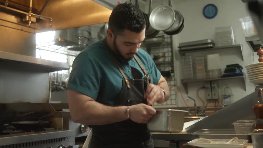 Man stirs small square metal bowl in restaurant kitchen