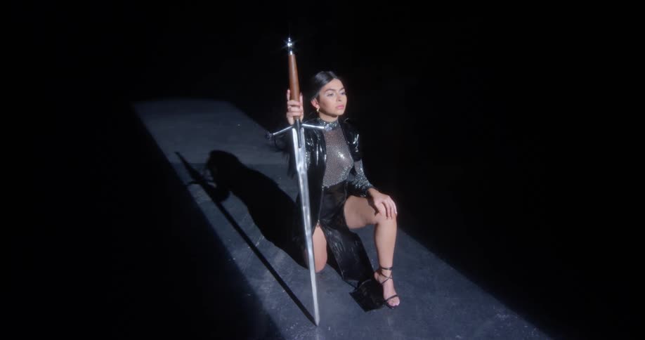 A woman is kneeling in the dark room holding a sword