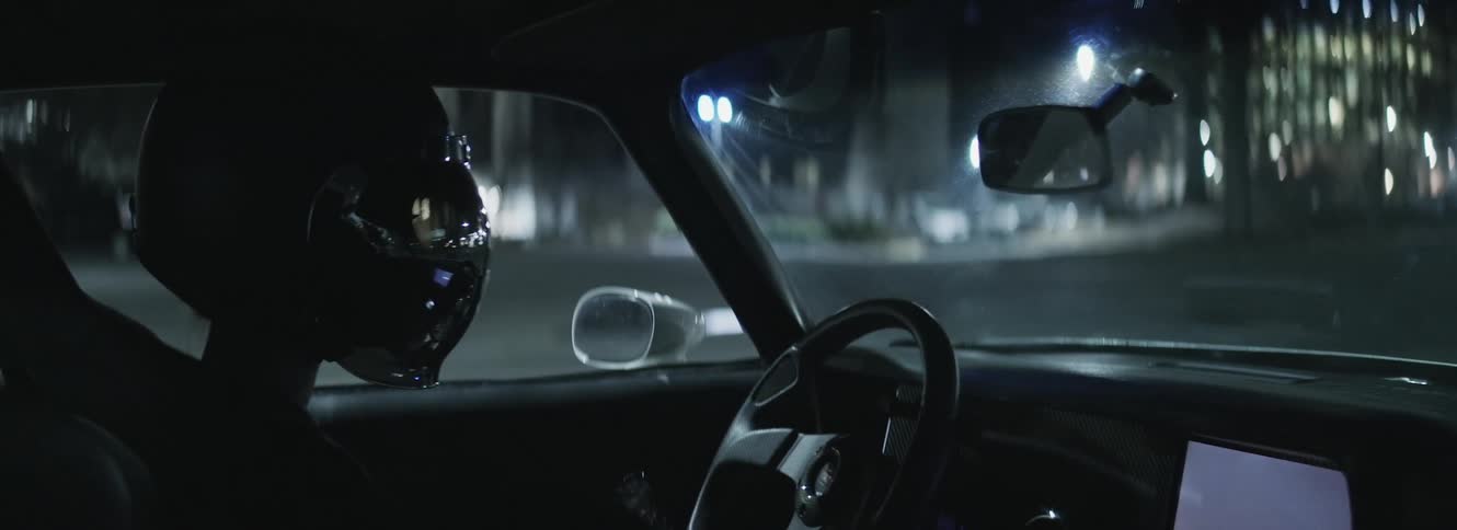 A driver wearing a helmet with black visor sits in a car looking at a screen display on the dash