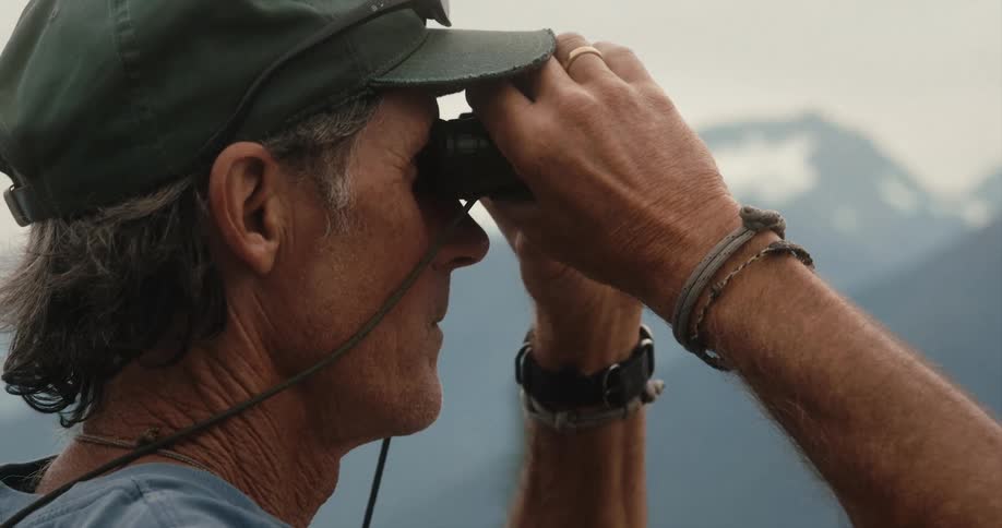 Man with sunglasses and green cap looks at mountains through binoculars