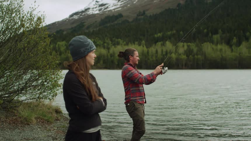 A woman stands with arms crossed near a man fishing in a mountain lake