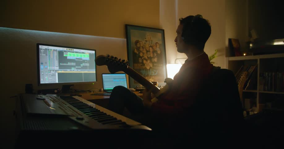 A male guitarist strums on his electric guitar while recording his music on a computer