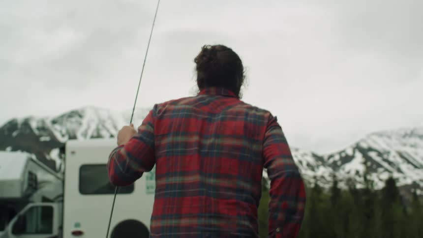 A person walking with a fishing rod with a mountain range the sky and clouds in the background