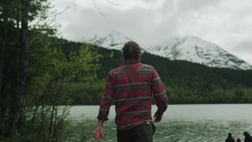A man in a plaid flannel shirt walks towards a large lake next to snow covered mountains